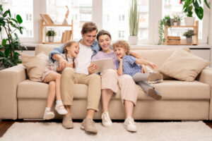 Young joyful casual family of two kids and couple sitting on sofa and watching funny video or cartoons in touchpad