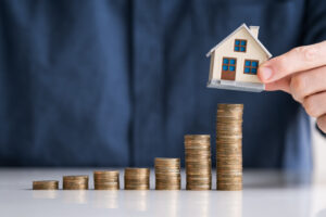 Close-up Hand Arranging House Model On Stacked Coins At Desk